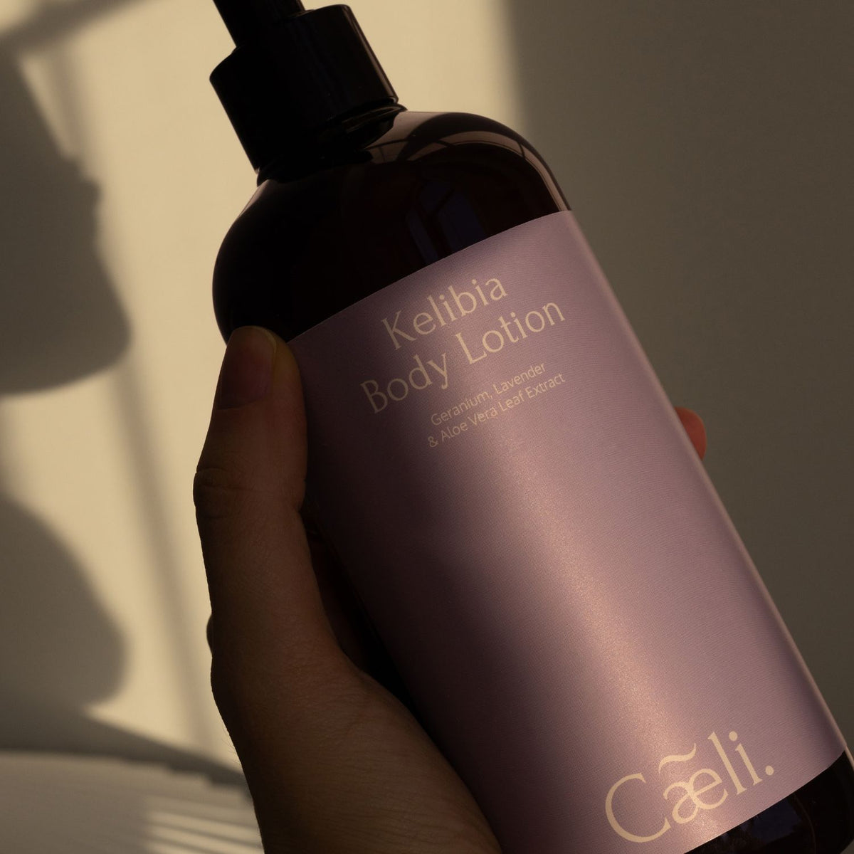 A hand holding a dark amber bottle of Kelbia Organic Body Lotion by Caeli. The bottle has a black pump and a dusty lavender-purple label with gold text, set against a warm, shadowed background.