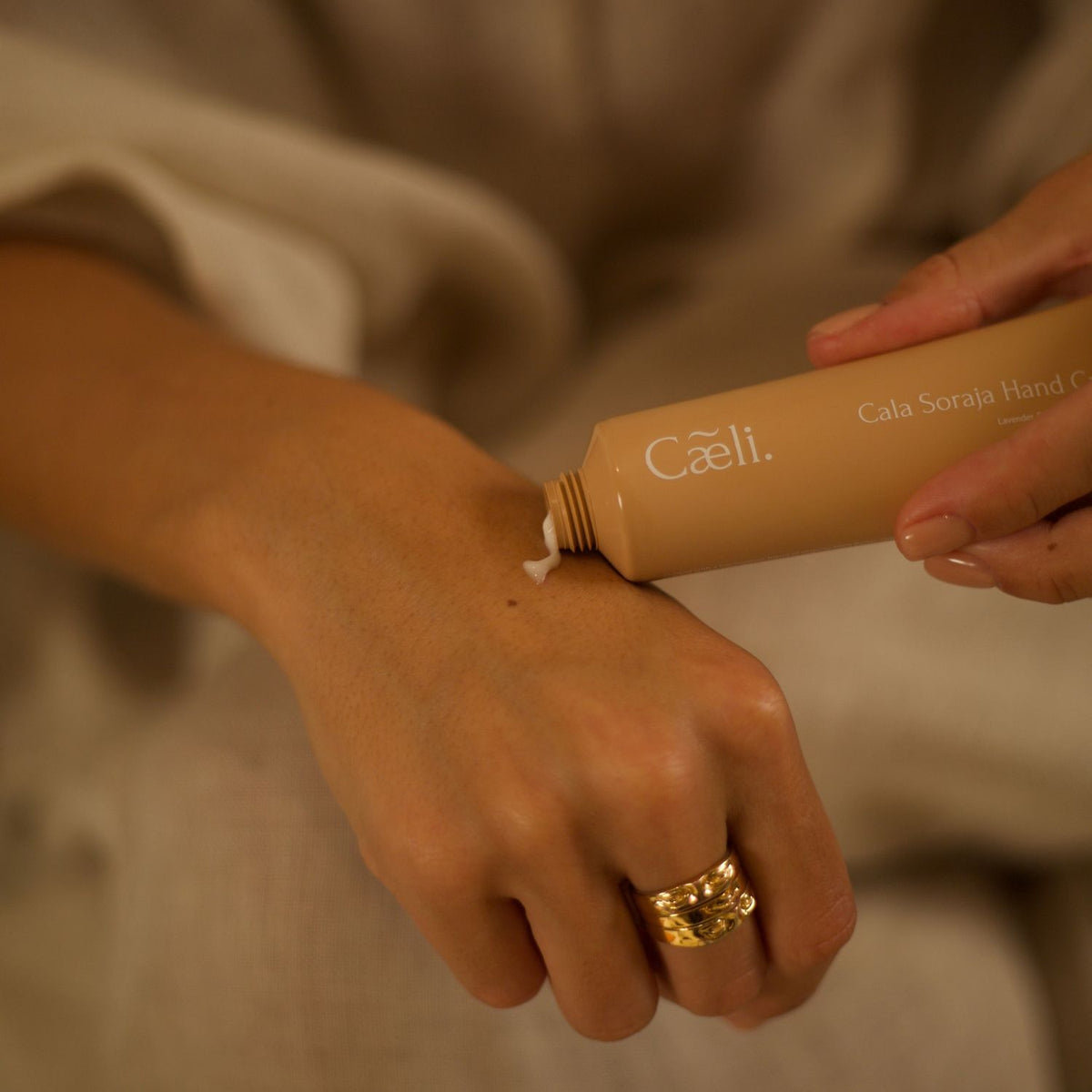 A close-up of a person's hands squeezing a beige cosmetic tube labeled 'Caeli Cala Soraja Hand cream' onto the back of another hand, with a warm neutral background.