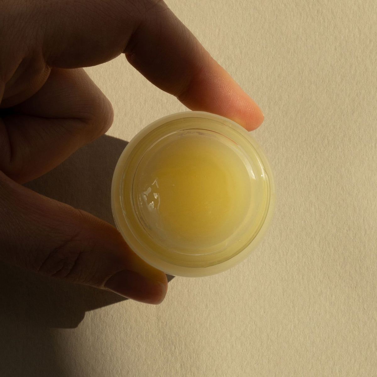 A hand holding a small round lip balm with a translucent pale yellow balm visible inside a clear container, against a plain beige background.