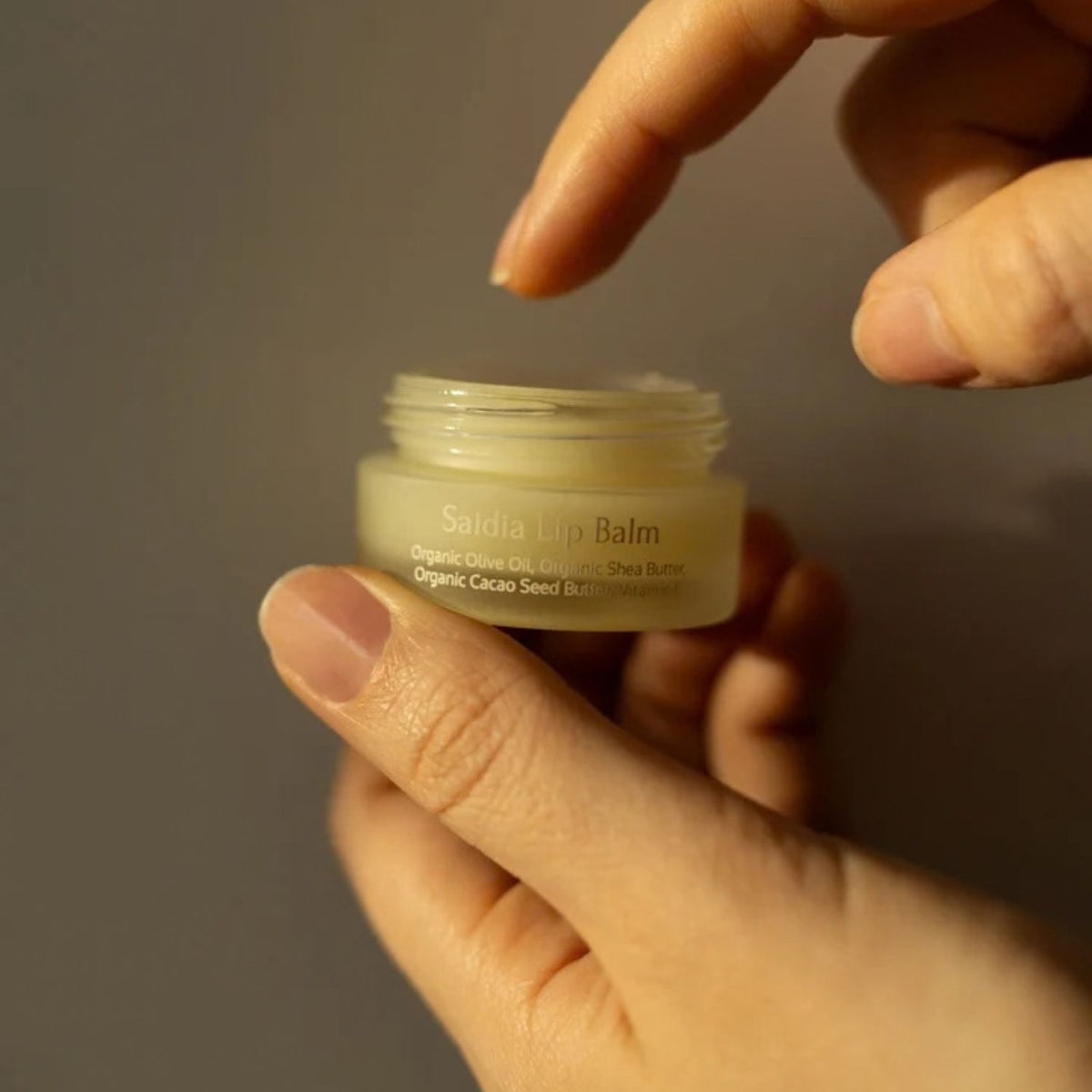 A hand holding an open jar of Caeli Saidia Lip Balm, with another hand pointing toward the jar, against a dark gray background.