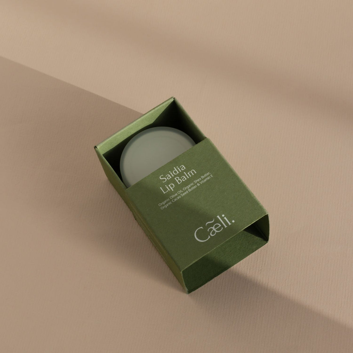 A square sage green box of Caeli Organic Lip Balm partially open, revealing a round pale green balm inside, set on a warm beige background with soft diagonal shadows.
