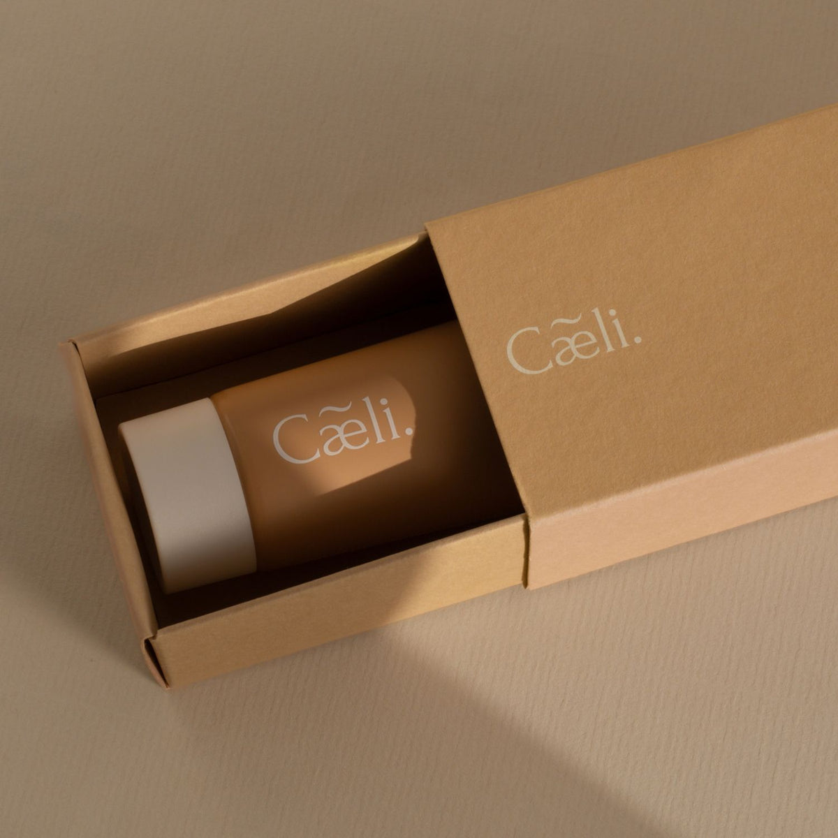A cylindrical cosmetic tube with a white cap and the brand name 'Caeli' printed on it, nestled inside an open kraft paper box.