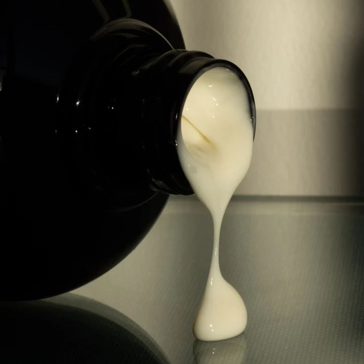 A close-up of a creamy body lotion dripping from a glossy amber bottle onto a reflective gray surface.