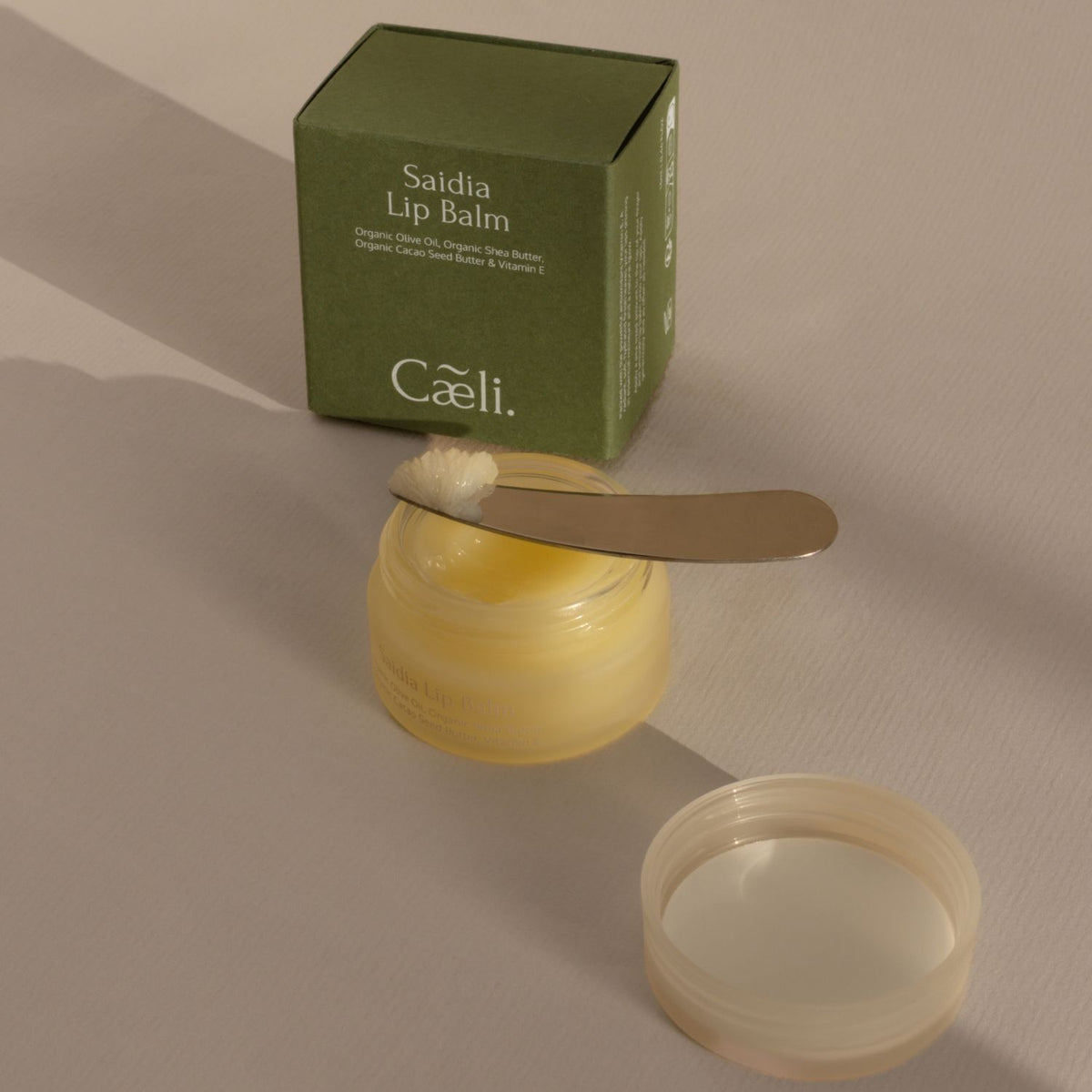 Caeli Saidia Lip Balm, with organic olive oil and shea butter, in a translucent yellow jar with its green box and a metal spatula with lip balm on it, on a neutral beige background.