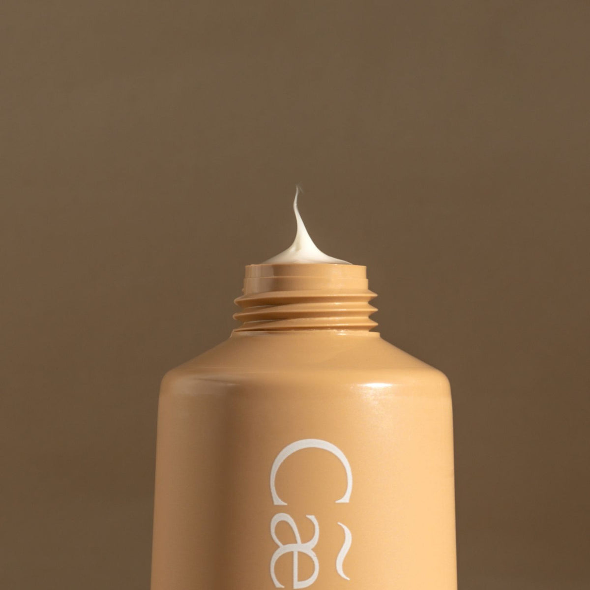 A close-up of a beige cosmetic tube with its cap removed, showing a dollop of white hand cream on top, against a solid taupe-brown background. The brand name 'Caeli' is partially visible in white script on the bottle.